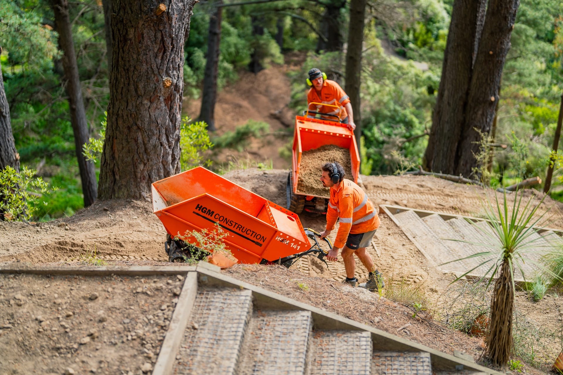 Civil contractors using power barrows to move material across a forest walking track during recreation construction and park construction works. The image highlights walking track construction, siteworks, erosion control, machinery transport and project management in difficult access terrain.