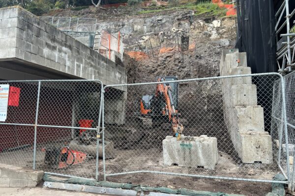 Retaining walls and civil construction underway on a steep coastal site, with excavation, slope retention and site preparation works taking place behind temporary fencing. The image shows civil contractors managing complex construction projects involving concrete structure works, ground stabilisation and rockfall mesh and fencing.