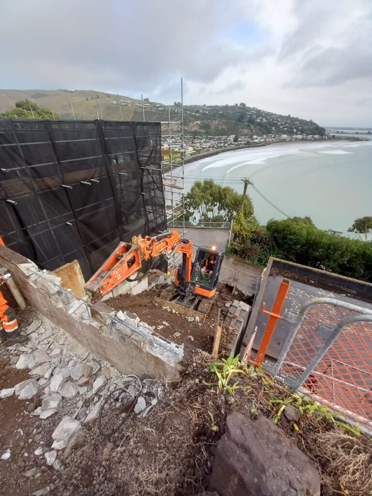 Excavation and retaining walls construction on a steep coastal site overlooking the shoreline, with civil contractors and machinery working beside scaffolding and safety fencing. This image highlights civil construction, siteworks, foundation construction and slope retention for a challenging residential project.