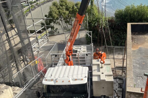 Crane truck positioned on a coastal construction site for lifting and sling operations during retaining walls and civil construction works. The image shows civil contractors managing machinery transport, site construction and project management beside scaffolding on a steep residential site.