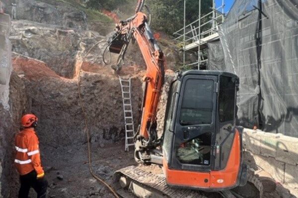 Civil contractors using excavation equipment and a construction drill to prepare a steep rock face for retaining walls, slope retention and ground stabilisation. The image shows drilling services, site preparation, rock anchors and soil nails being used as part of complex construction projects.