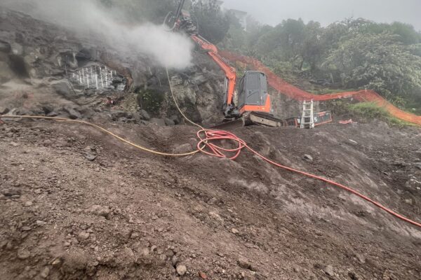 Excavation and drilling services on a steep hillside, with a construction drill working through rock during civil construction and siteworks. This image shows civil contractors carrying out slope retention, rock anchors, soil nails and ground stabilisation for a challenging retaining walls project.