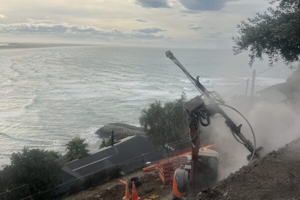 Civil contractors using drilling services on a steep coastal construction site, with excavation equipment working above the shoreline during retaining walls and slope retention works. This image shows site construction, ground stabilisation, rock anchors and complex project management in a difficult hillside location.