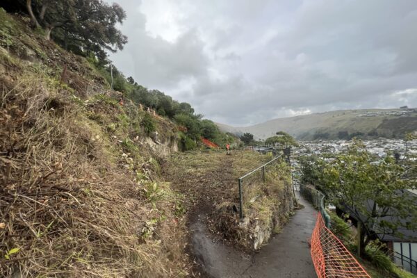 Site preparation and excavation along a narrow hillside access path, with safety fencing and coastal views beside the retaining walls construction area. The image shows civil contractors managing slope retention, ground stabilisation and civil construction works on a constrained steep site.