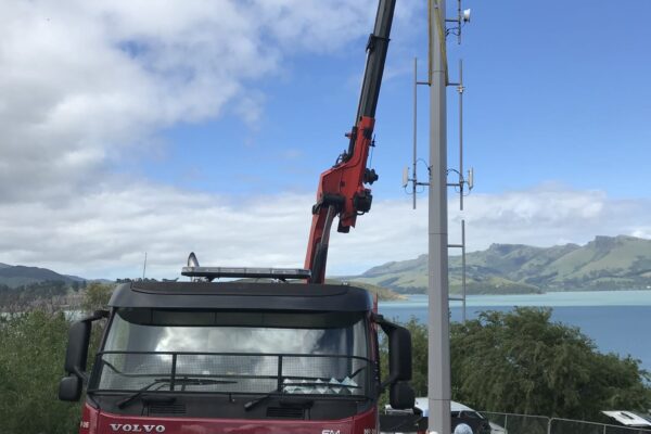 Crane truck lifting a tower pole during AIS tower installation on a coastal site, with civil contractors managing utility infrastructure and overhead infrastructure works. The image highlights civil construction, machinery transport, sling operations and project management in a difficult access location.
