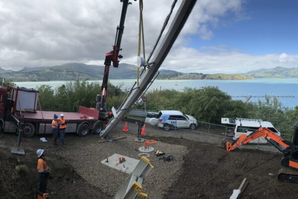 Panoramic view of crane and excavation equipment being used for AIS tower installation and utility infrastructure works above the harbour. The image shows civil contractors completing site preparation, machinery transport, sling operations and tower installation in a remote coastal setting.