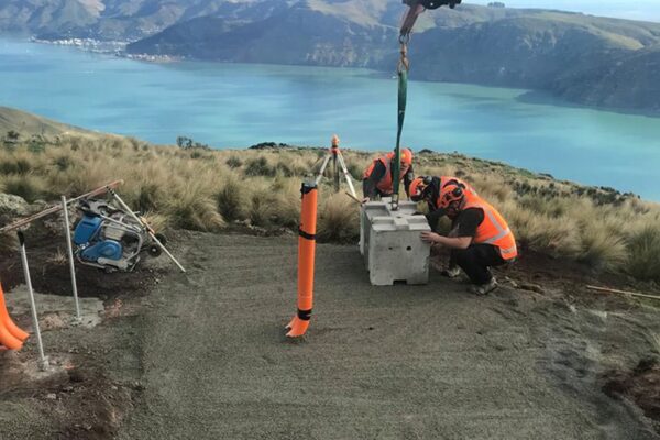 Civil contractors positioning a concrete foundation block during AIS tower installation on a remote coastal hillside, with crane lifting equipment visible above. The image shows utility infrastructure, tower installation, sling operations, foundation construction and civil construction siteworks.