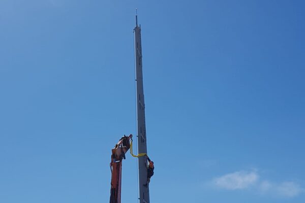 Excavator positioning a tall tower pole during AIS tower installation on a rocky coastal hilltop. The image highlights civil construction, tower installation, utility infrastructure, overhead infrastructure and project management in a difficult access location.