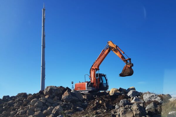 Excavator carrying out siteworks around a newly installed tower pole on a rocky hilltop site. The image shows civil contractors delivering tower installation, utility infrastructure, foundation construction and site preparation for overhead infrastructure in a remote location.