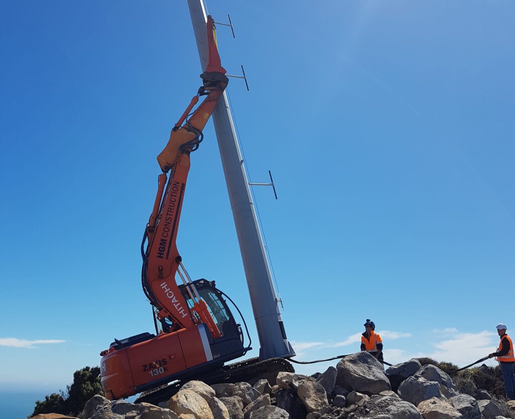 Excavator assisting with tower installation on a rocky coastal hilltop, with civil contractors guiding the tall pole into place. This image highlights civil construction, utility infrastructure, overhead infrastructure, foundation construction and project management in difficult access terrain.