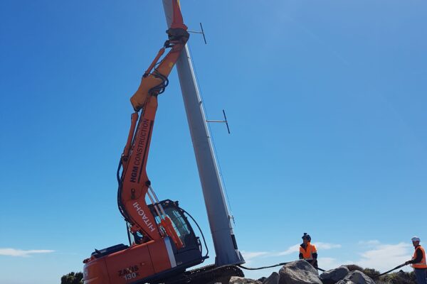 Excavator assisting with tower installation on a rocky coastal hilltop, with civil contractors guiding the tall pole into place. This image highlights civil construction, utility infrastructure, overhead infrastructure, foundation construction and project management in difficult access terrain.