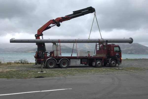 Crane truck transporting and lifting a tower pole for AIS tower installation beside a coastal road. The image shows civil contractors managing machinery transport, sling operations, utility infrastructure and overhead infrastructure works.