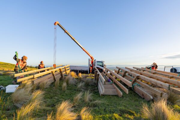 Civil contractors preparing timber materials for a staircase replacement project at Barnett Park, using crane equipment and sling operations in a coastal hill setting. The image highlights recreation construction, walking track construction, timber structures, machinery transport and project management in difficult access terrain.