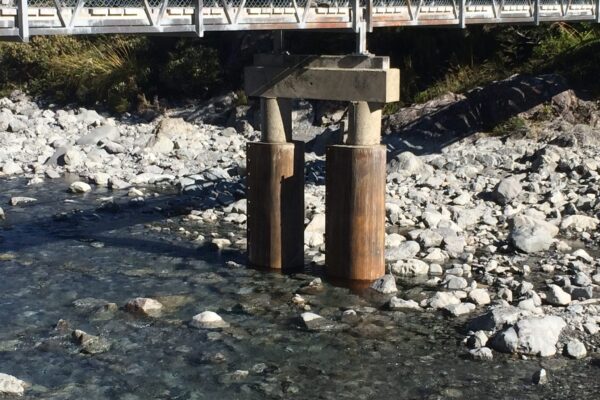 Bridge repair and bridge inspection works around timber piles and a concrete bridge support in a rocky riverbed. The image shows civil contractors completing bridge maintenance, piling contractors, driven piles and siteworks beneath an existing bridge structure.