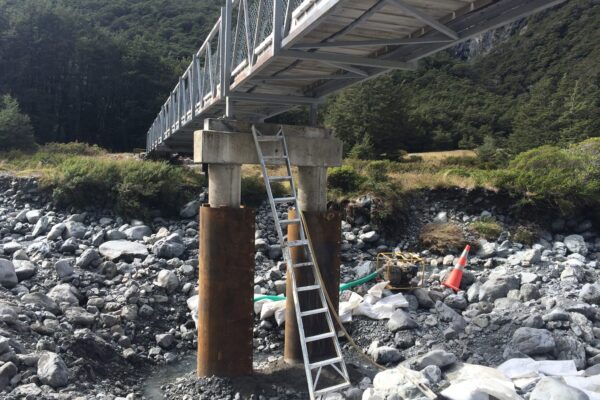 Bridge repair works taking place beneath an existing bridge, with ladders and equipment set up around timber piles and concrete supports. The image shows civil contractors carrying out bridge maintenance, bridge inspection, piling works and site preparation in a rocky riverbed.