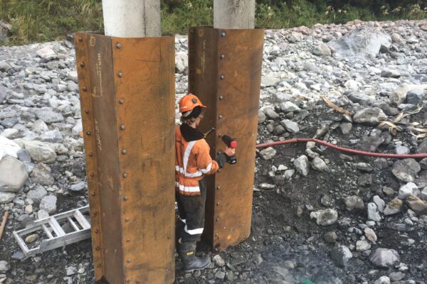Civil contractor working between timber piles beneath a bridge during bridge repair and bridge strengthening works. The image highlights bridge maintenance, bridge inspection services, piling contractors, driven piles and civil construction in a rocky waterway.