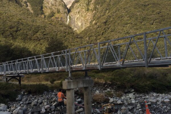 Existing bridge structure crossing a rocky riverbed, with civil contractors working below on bridge repair and bridge inspection services. The image highlights bridge maintenance, bridge strengthening, piling contractors, driven piles and project management in a remote alpine setting.