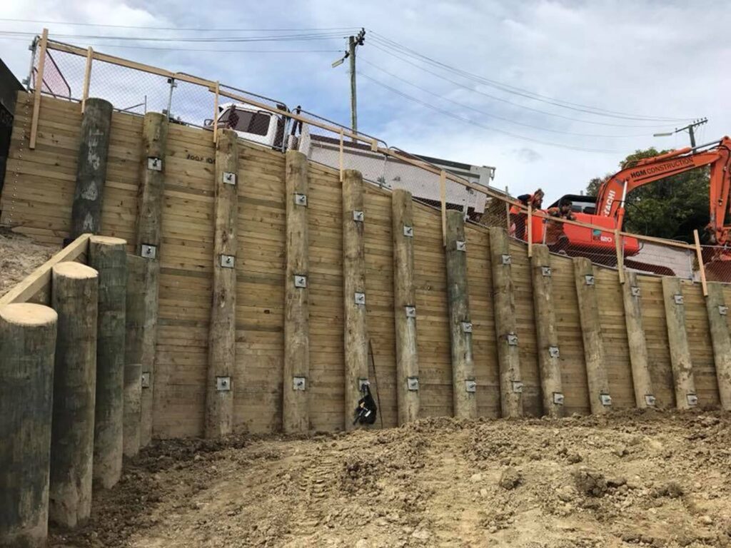 Large scale timber retaining walls completed on a steep site, with mechanical anchors, excavation equipment and civil contractors visible at the top of the structure. This image shows civil construction, slope retention, ground stabilisation, foundation construction and complex project management.