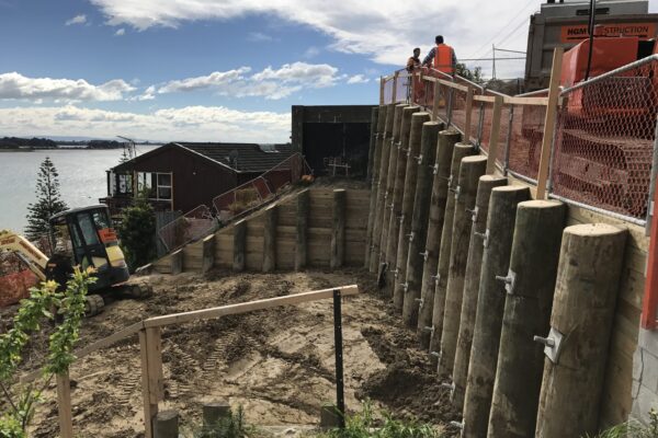 Large scale retaining walls built with timber poles on a steep coastal site, with civil contractors completing excavation and site preparation above nearby homes. The image highlights retaining walls, slope retention, ground stabilisation, foundation construction and complex project management.