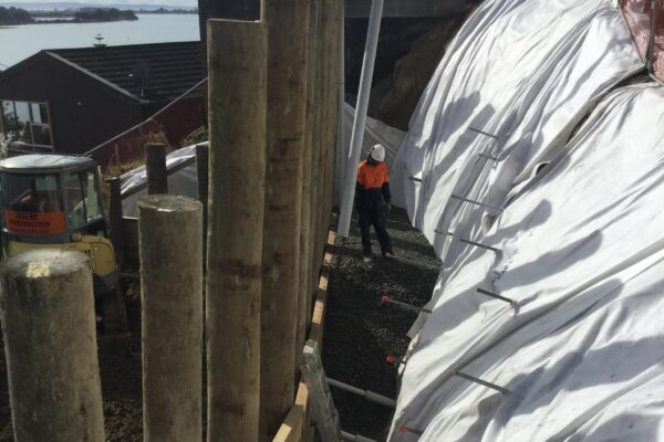 Large scale retaining walls under construction on a steep coastal site, with timber piles installed beside a covered slope and nearby residential buildings. This image highlights civil construction, slope retention, ground stabilisation and site preparation by civil contractors.