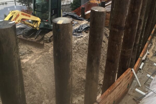 Timber pile retaining walls being built above a coastal residential area, with excavation machinery working below the structure. The image shows civil contractors managing foundation construction, driven piles, slope retention and complex construction projects in a constrained site.