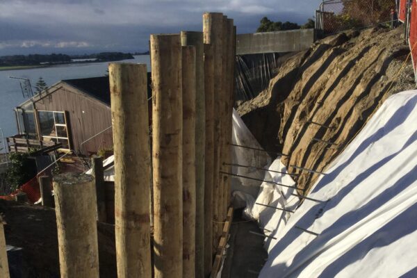 Timber retaining walls and slope protection works on a steep site overlooking the coast, with covered earthworks and installed piles visible along the excavation. This image shows civil construction, retaining walls, ground stabilisation, site preparation and project management by civil contractors.