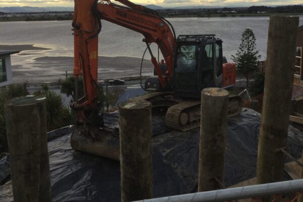 Excavation machinery working among timber piles during retaining walls construction on a coastal hillside site. The image highlights civil construction, siteworks, earthworks contractors, foundation construction and slope retention for a large scale retaining walls project.