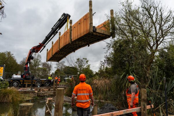 Timber bridge section suspended by crane above a reserve waterway during installation by civil contractors. The image highlights bridge construction, sling operations, timber structures and site construction for recreation acc