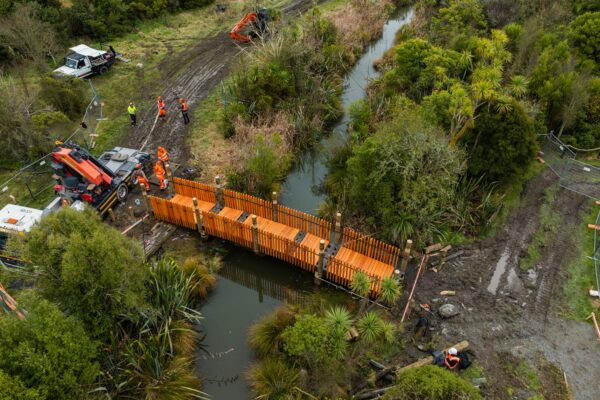 Aerial view of civil construction crews installing a timber bridge over a waterway, with excavation equipment and siteworks visible around the bridge construction area. This image shows civil contractors managing complex construction projects involving timber structures, erosion control, park construction and walking track construction.