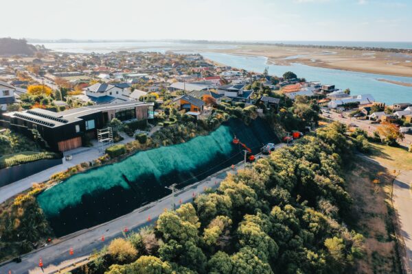 Aerial view of large scale slope retention and erosion control works beside a coastal road and residential area. The image shows civil construction, ground stabilisation, rockfall mesh and fencing, and siteworks completed by experienced civil contractors.