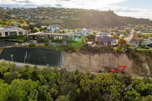 Wide aerial view of coastal erosion control works, with an excavator operating below a steep hillside near residential properties. This civil construction project includes slope retention, ground stabilisation, rockfall mesh and fencing, and careful project management.