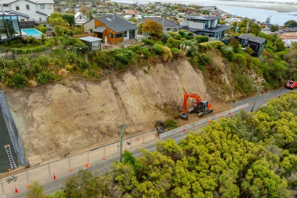Excavator completing site preparation and slope retention works along a coastal road below residential properties. The image highlights civil construction, erosion control, ground stabilisation and excavation work on a steep roadside site.