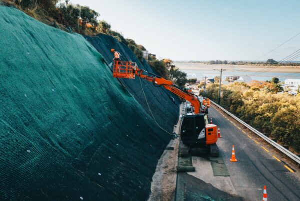 Excavator and rope access worker installing rockfall mesh and fencing across a steep roadside slope for erosion control. This civil construction project includes slope retention, ground stabilisation, site preparation and work on a restricted coastal site.