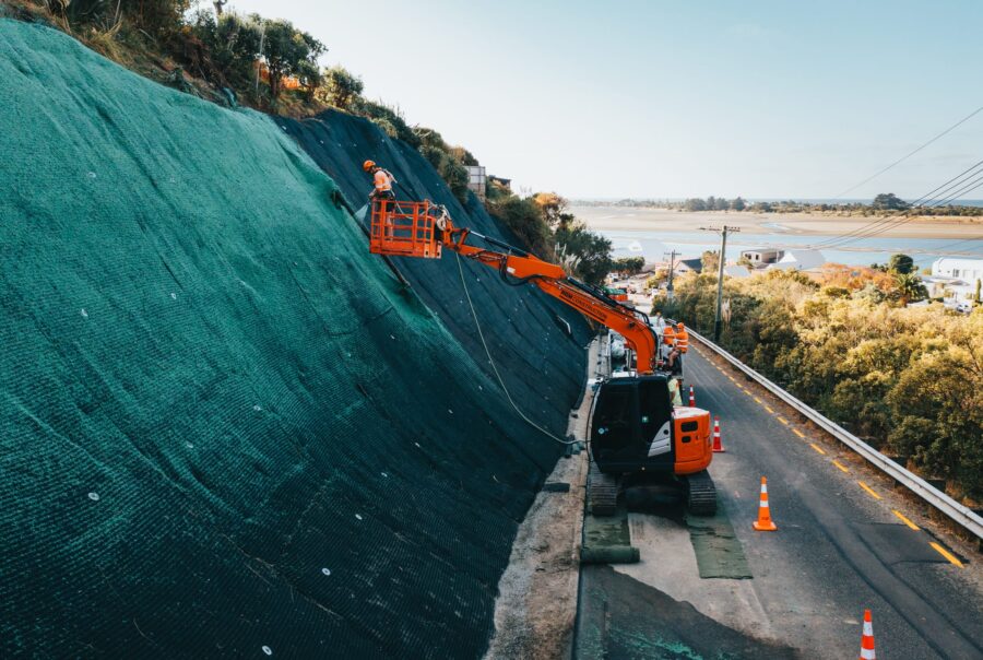 Excavator and rope access worker installing rockfall mesh and fencing across a steep roadside slope for erosion control. This civil construction project includes slope retention, ground stabilisation, site preparation and work on a restricted coastal site.