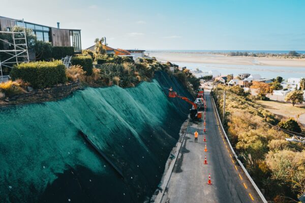 Steep coastal roadside slope covered with rockfall mesh and fencing during erosion control and slope retention works. The image demonstrates civil construction, ground stabilisation, siteworks and careful project management near residential properties.