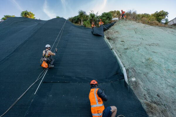Rope access workers installing rockfall mesh across a steep slope as part of erosion control and civil construction works. The project shows slope retention, ground stabilisation, site preparation and complex construction work on an exposed hillside.