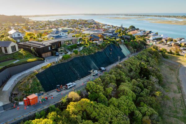 Aerial view of erosion control and slope retention works protecting a steep hillside road near residential properties and the coastline. This civil construction project includes rockfall mesh and fencing, site preparation and ground stabilisation.