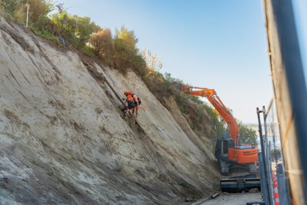 Excavator and rope access worker carrying out slope retention and erosion control on a steep roadside bank. The image shows civil construction, excavation, rockfall mesh installation and site preparation in a restricted coastal environment.