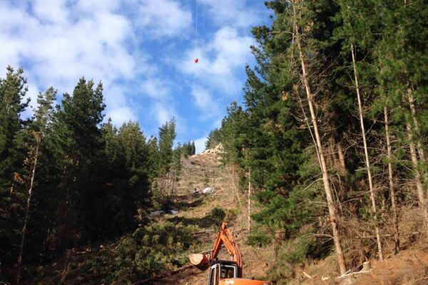 Helicopter operation supporting civil construction in a steep forest corridor, with excavation machinery and site contractors working below. This image shows commercial sling operations, adventure infrastructure, park construction and complex project management in difficult terrain.