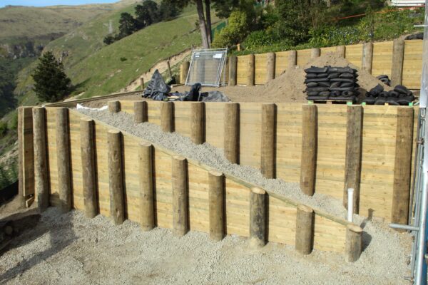 Timber retaining walls with driven piles and gravel backfill forming a stepped slope retention structure. The image shows civil construction, foundation construction, ground stabilisation and site preparation on a steep residential development.