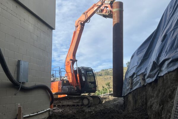 Excavator installing a driven pile for timber retaining walls on a confined residential site. The image shows civil construction, excavation, slope retention, foundation construction and site preparation by experienced civil contractors in New Zealand.