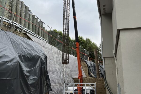 Truck-mounted crane carrying out sling operations to position construction drill equipment for retaining walls and piling work. The image shows civil construction, drilling services, foundation construction and complex project management on a confined residential site.