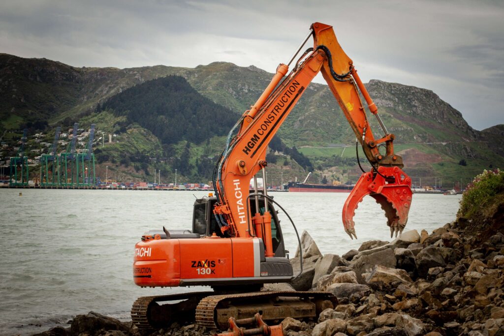 Excavator completing marine construction and coastal construction works along a rocky shoreline, using a grapple attachment for excavation and dredging near the water. This image highlights marine services, marine contractors, civil contractors and site construction for utility infrastructure.