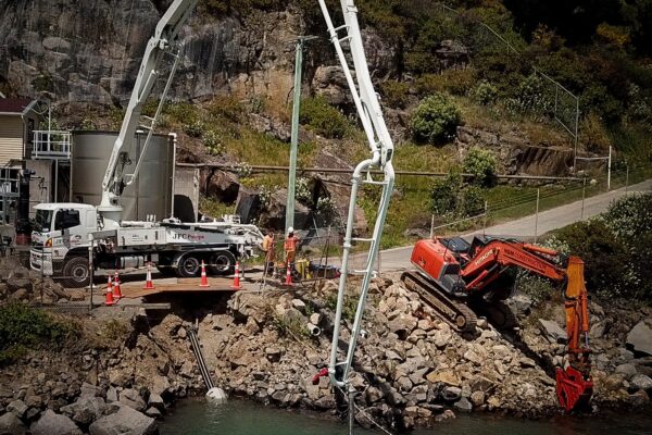 Marine civil contractors completing pipeline and utility infrastructure works on a rocky shoreline, with concrete pumping equipment and excavation machinery working beside the harbour. The image shows marine construction, coastal construction, marine services and complex civil construction project management.