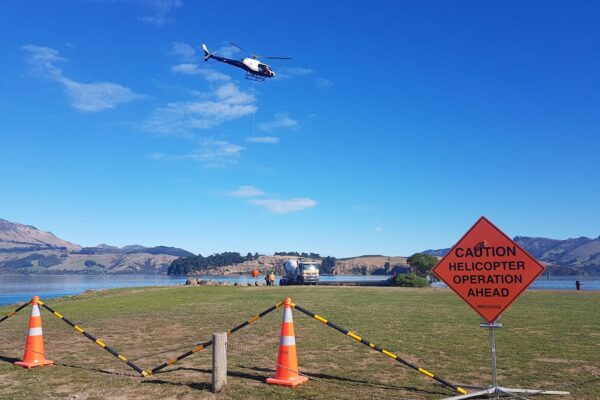Helicopter operation taking place near a coastal worksite on Quail Island, with safety cones and signage marking the helicopter operation area. The image shows civil contractors managing heli operation, commercial sling logistics, site preparation and project management for remote island construction.