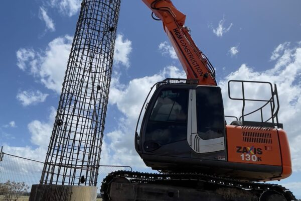 Excavator lifting a reinforcing cage into position for a bored pile foundation during power infrastructure construction. The image shows civil contractors managing foundation construction, anchoring and piling, concrete structure preparation and transmission line construction.