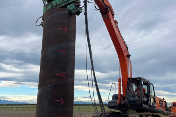 Excavation equipment lifting a large steel casing during bored pile foundation construction for power infrastructure. The image shows civil contractors completing anchoring and piling, siteworks, utility infrastructure and ground stabilisation for a transmission line construction project.