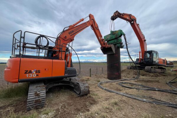 Civil contractors using excavation machinery to position a steel casing during foundation construction for overhead infrastructure. This image highlights bored piles, anchoring and piling, power infrastructure, site preparation and project management on a rural construction site.
