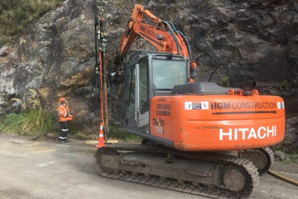 Excavator-mounted construction drill working beside a rocky roadside slope during civil construction and drilling services. The image shows civil contractors carrying out rock anchors, soil nails, slope retention and ground stabilisation on a constrained road site.