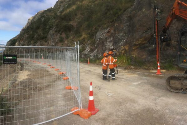Wide view of civil construction on a narrow coastal road, with excavation equipment, safety fencing and civil contractors preparing for slope retention works. The image highlights site preparation, rock anchors, ground stabilisation and project management on a complex hillside construction site.